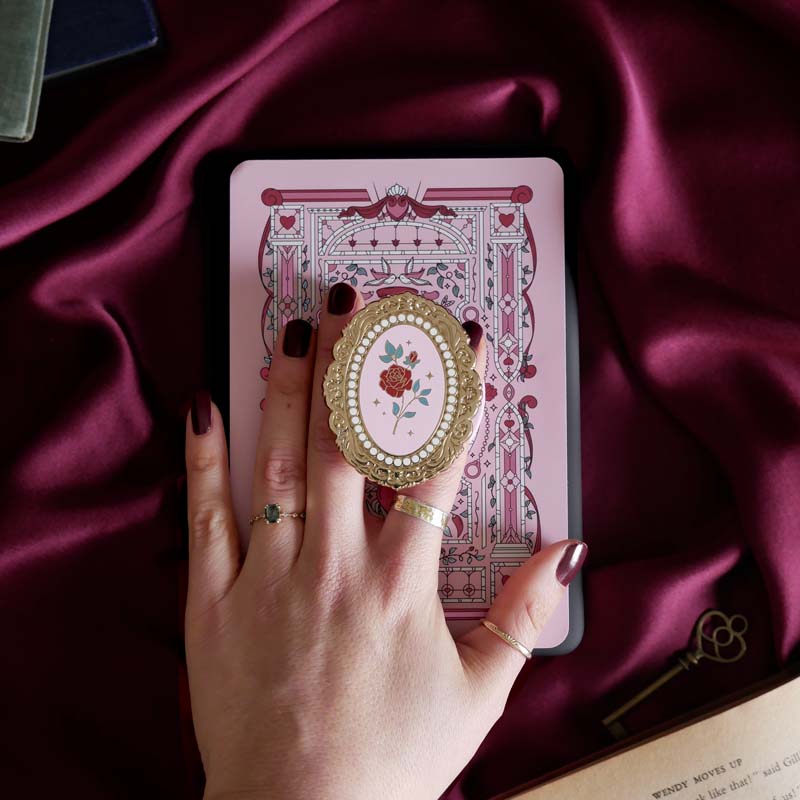 Hand holding a decorative kindle grip with a pink coquette rose inspired design with gold accents on a pink fabric background