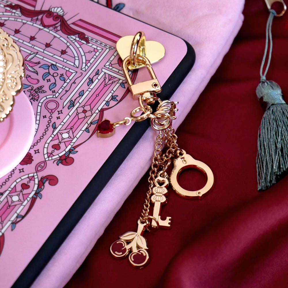 Sugar & Spice eReader bundle image for romance book kindle decor- a gold charmlet with cherries, keys, a handcuff and gem is attached to an eReader with matching pink eReader sticker insert. The scene sits on a pink eReader case on red fabric.
