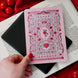 A hand holding a Kindle sticker insert over an e-reader on a background of a book and red fabric. The eReader sticker insert is light pink and features an intricate design with roses, birds, flowers, columns, hands and handcuff motifs in shades of red, pink and green.