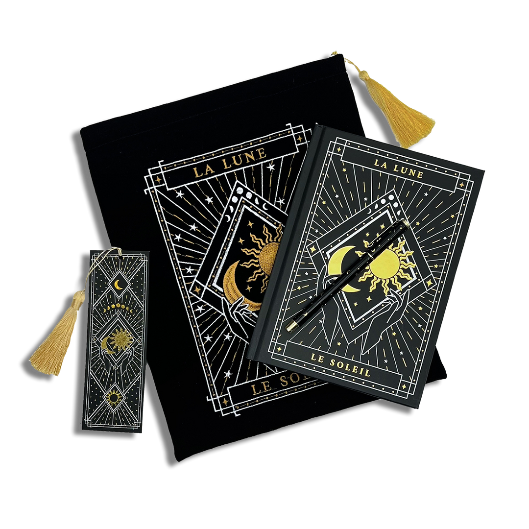 bundle of black stationery items on a plain background. Items are a book sleeve, journal, pen and bookmark. Design features a moon and sun caressed by hands with celestial inspired design. 
