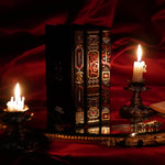 On a red fabric background sits the three journals for the Serpents & Swords collection- the journal, reading journal and reading companion. Sitting on a tray with two lit wax candles to the side, the journals are all black and feature intricate red and gold foiling that show motifs of snakes, swords, dragons, crowns, goblets and more from a romantasy book aesthetic.