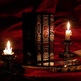 On a red fabric background sits the three journals for the Serpents & Swords collection- the journal, reading journal and reading companion. Sitting on a tray with two lit wax candles to the side, the journals are all black and feature intricate red and gold foiling that show motifs of snakes, swords, dragons, crowns, goblets and more from a romantasy book aesthetic.