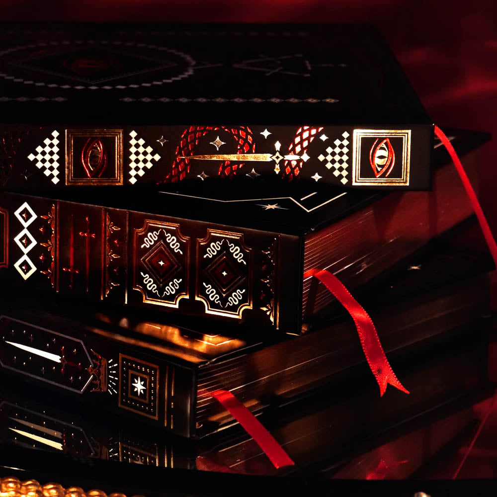 A detail shot of The Swords & Serpents journals. On a red fabric background sits the three journals- the journal, reading journal and reading companion. Stacked on their side, the journals are all black and feature intricate red and gold foiling that show motifs of snakes, swords, dragons, crowns, goblets and more from a romantasy book aesthetic.