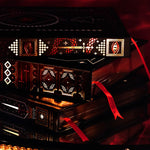 A detail shot of The Swords & Serpents journals. On a red fabric background sits the three journals- the journal, reading journal and reading companion. Stacked on their side, the journals are all black and feature intricate red and gold foiling that show motifs of snakes, swords, dragons, crowns, goblets and more from a romantasy book aesthetic.