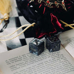A black Quirky Cup Collective loot pouch with red interior and gold chord detailing sits on an open book and as chessboard, two black dice in the foreground.