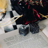 A black Quirky Cup Collective loot pouch with red interior and gold chord detailing sits on an open book and as chessboard, two black dice in the foreground.