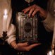 "A person in a vintage-style blouse holds a Black Reading Journal by The Quirky Cup Collective, showcasing its ornate gold and white embossed cover with the words 'Reading Journal' in the center. The scene is dimly lit by candlelight, creating a warm, mysterious ambiance. The journal is framed by elegant candelabras with lit candles, and a glass of red wine is seen in the foreground, enhancing the moody and intimate setting