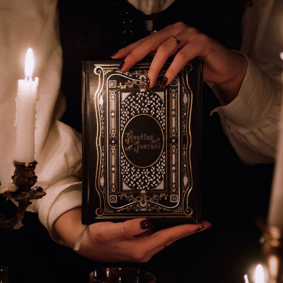 "A person in a vintage-style blouse holds a Black Reading Journal by The Quirky Cup Collective, showcasing its ornate gold and white embossed cover with the words 'Reading Journal' in the center. The scene is dimly lit by candlelight, creating a warm, mysterious ambiance. The journal is framed by elegant candelabras with lit candles, and a glass of red wine is seen in the foreground, enhancing the moody and intimate setting