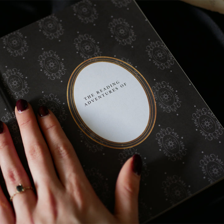 A hand with dark red nail polish and a delicate ring rests on the cover of a Black Reading Journal by The Quirky Cup Collective, showcasing its ornate gold and white embossed design and the words 'Reading Journal' in the center. The journal lies on a smooth black satin fabric creating a luxurious and elegant ambiance