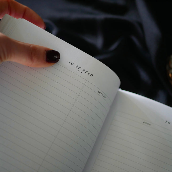 A hand with dark red nail polish and a delicate ring rests on the cover of a Black Reading Journal by The Quirky Cup Collective, showcasing its ornate gold and white embossed design and the words 'Reading Journal' in the center. The journal lies on a smooth black satin fabric creating a luxurious and elegant ambiance