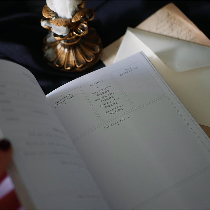 A hand with dark red nail polish and a delicate ring rests on the cover of a Black Reading Journal by The Quirky Cup Collective, showcasing its ornate gold and white embossed design and the words 'Reading Journal' in the center. The journal lies on a smooth black satin fabric, with a decorative candle holder nearby, creating a luxurious and elegant ambiance