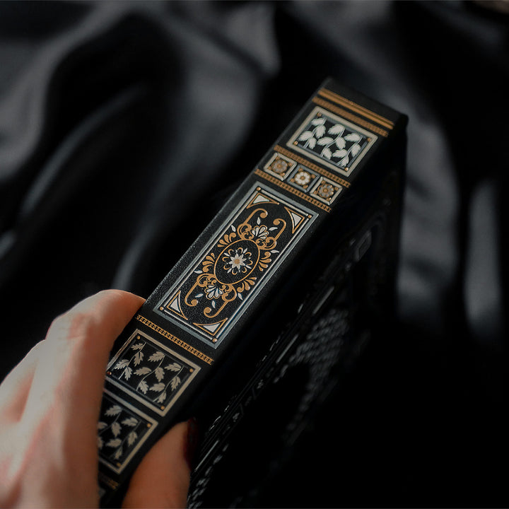 A hand with dark red nail polish and a delicate ring rests on the cover of a Black Reading Journal by The Quirky Cup Collective, showcasing its ornate gold and white embossed design and the words 'Reading Journal' in the center. The journal lies on a smooth black satin fabric creating a luxurious and elegant ambiance