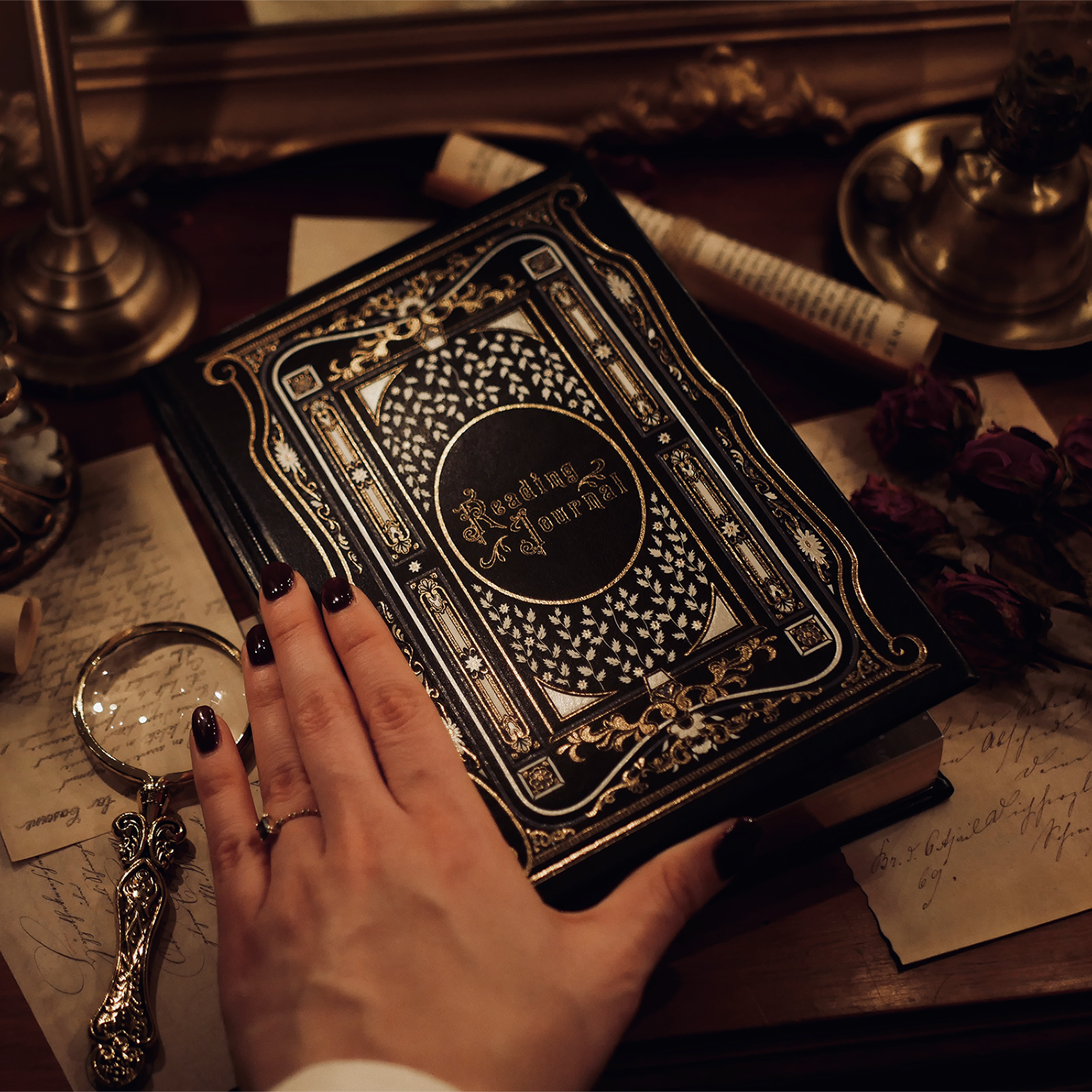 A hand with dark nail polish gently touching the cover of a Black Reading Journal by The Quirky Cup Collective, featuring intricate gold and white embossed designs and the words 'Reading Journal' in the center. The journal is placed on a vintage wooden desk surrounded by handwritten letters, a magnifying glass, dried roses, and a lit candle, creating an intimate, cozy atmosphere
