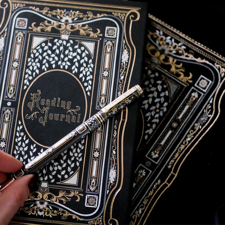 A hand holding a luxurious reading journal with an intricate gold and white design, accompanied by an ornate matching pen. The journal lies on top of a black velvet book sleeve from The Quirky Cup Collective, showcasing a coordinated design. A matching bookmark with a silver tassel is also partially visible. All items are set against a smooth black satin fabric background, creating an elegant and cohesive visual presentation.