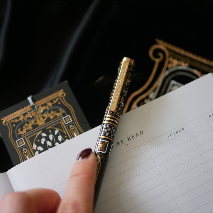 A hand holding a luxurious reading journal with an intricate gold and white design, accompanied by an ornate matching pen. The journal lies on top of a black velvet book sleeve from The Quirky Cup Collective, showcasing a coordinated design. A matching bookmark with a silver tassel is also partially visible. All items are set against a smooth black satin fabric background, creating an elegant and cohesive visual presentation.
