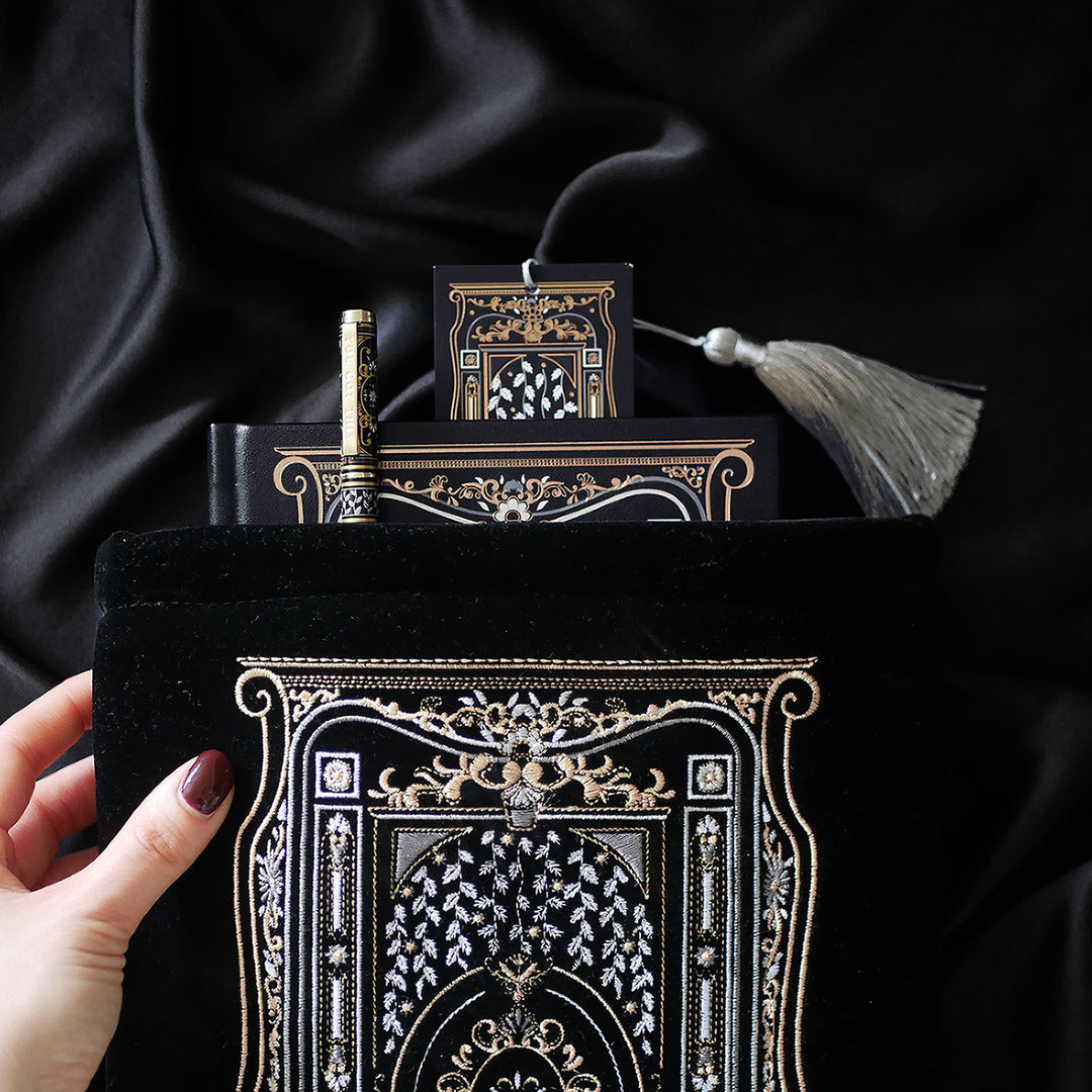 A hand holding a luxurious reading journal with an intricate gold and white design, accompanied by an ornate matching pen. The journal lies on top of a black velvet book sleeve from The Quirky Cup Collective, showcasing a coordinated design. A matching bookmark with a silver tassel is also partially visible. All items are set against a smooth black satin fabric background, creating an elegant and cohesive visual presentation.