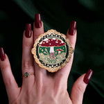 The gold wonderland clingie (similar to a book inspired pop socket!) features an intricate gold frame design with two little red mushrooms surrounded by plants in shades of green. The clingie is held in a hand with red nails in front of a green fabric background.