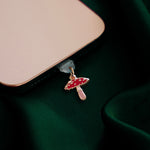 The wonderland enamel mushroom dust plug by The Quirky Cup collective is inserted into a gold iphone sitting on a dark green fabric background.