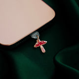 The wonderland enamel mushroom dust plug by The Quirky Cup collective is inserted into a gold iphone sitting on a dark green fabric background.