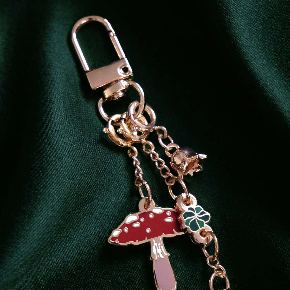 A detail of the  gold Wonderland charmlet by The Quirky cup collective sits on a green fabric background. The charm features multiple other charms including a small enamel red mushroom, a chain of green leaves and a small gold flower.