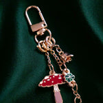 A detail of the  gold Wonderland charmlet by The Quirky cup collective sits on a green fabric background. The charm features multiple other charms including a small enamel red mushroom, a chain of green leaves and a small gold flower.