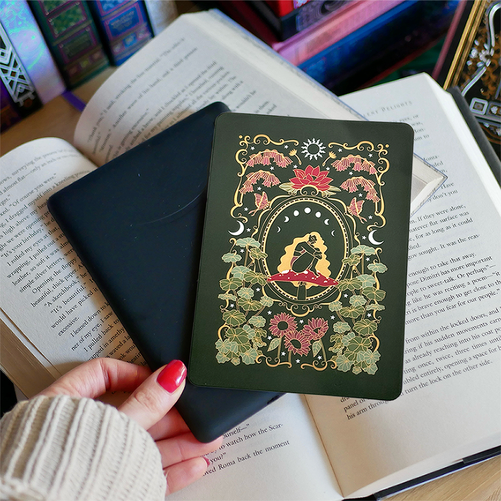 Image shows a hand gently touching a kindle and a kindle sticker insert sitting on top of the device. They sit on a bed of open books. The cottage core design has the colours green, red, gold and white. It shows a decorative frame showing a girl sitting on a mushroom. She is surrounded by narsurtium leaves, plants, flowers, butterflies and celestial embellishments