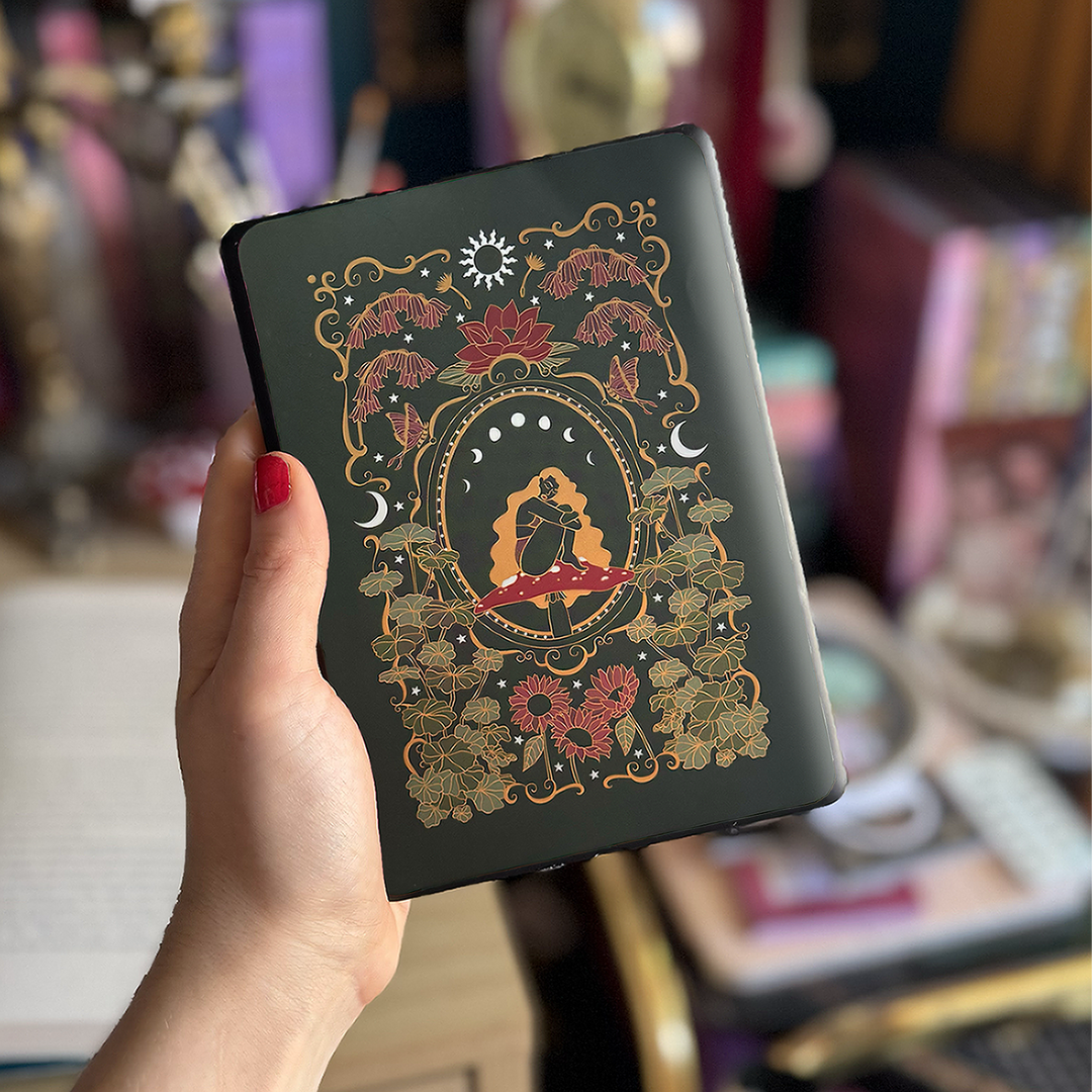 Image shows a hand holding a kindle showing off a kindle sticker insert that is placed on the back of the device. The cottage core design has the colours green, red, gold and white. It shows a decorative frame showing a girl sitting on a mushroom. She is surrounded by narsurtium leaves, plants, flowers, butterflies and celestial embellishments 