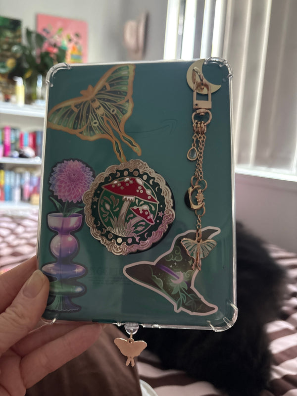Celestial inspired stickers, a luna moth charmlet and a mushroom phone grip are attached to a kindle