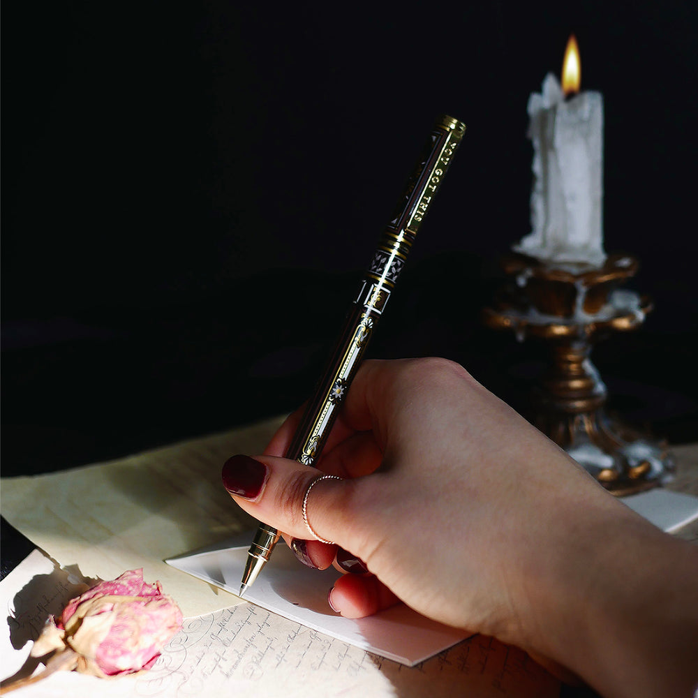 A hand holding the Black You Got This Pen by The Quirky Cup Collective, showcasing its intricate gold and white floral and geometric barrel design on a black background. The pen is positioned on top of a dark, silky fabric, with dried pink roses placed on top of a group of handwritten letters, creating a moody, vintage-inspired scene. 