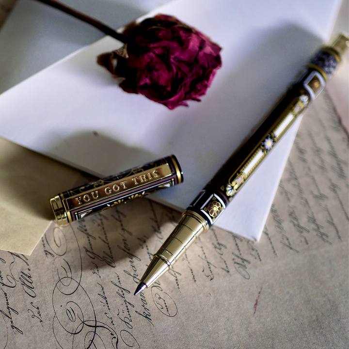 A hand holding the Black You Got This Pen by The Quirky Cup Collective, showcasing its intricate gold and white floral and geometric barrel design on a black background. The pen is positioned on top of a group of letters and envelopes with dried red roses creating a moody, vintage-inspired scene. 