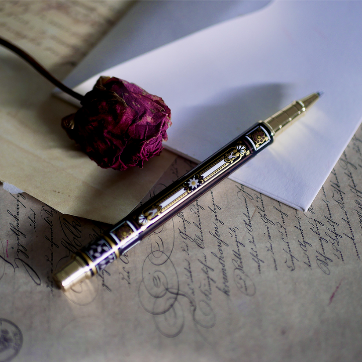 A hand holding the Black You Got This Pen by The Quirky Cup Collective, showcasing its intricate gold and white floral and geometric barrel design on a black background. The pen is positioned on top of a group of letters and envelopes with dried red roses creating a moody, vintage-inspired scene. 