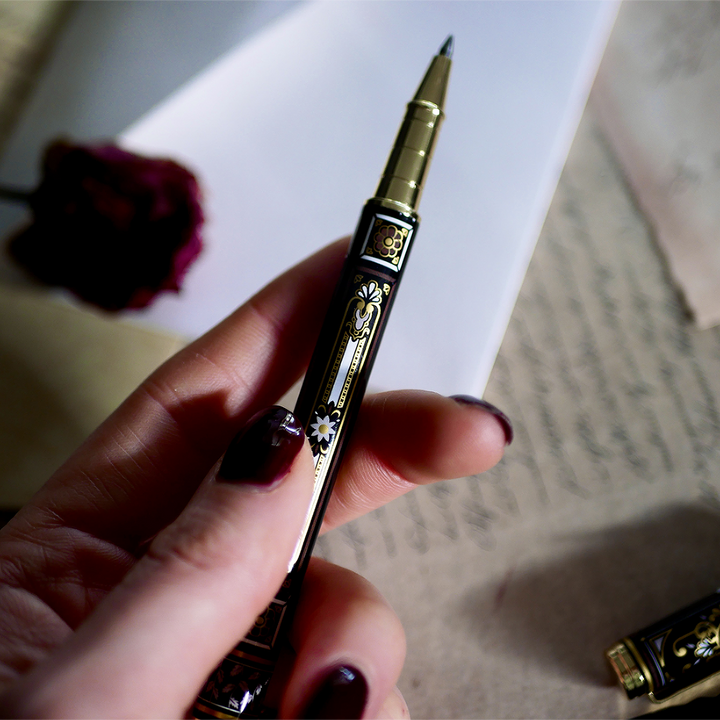 A hand holding the Black You Got This Pen by The Quirky Cup Collective, showcasing its intricate gold and white floral and geometric barrel design on a black background. The pen is positioned on top of a group of letters and envelopes with dried red roses creating a moody, vintage-inspired scene. 