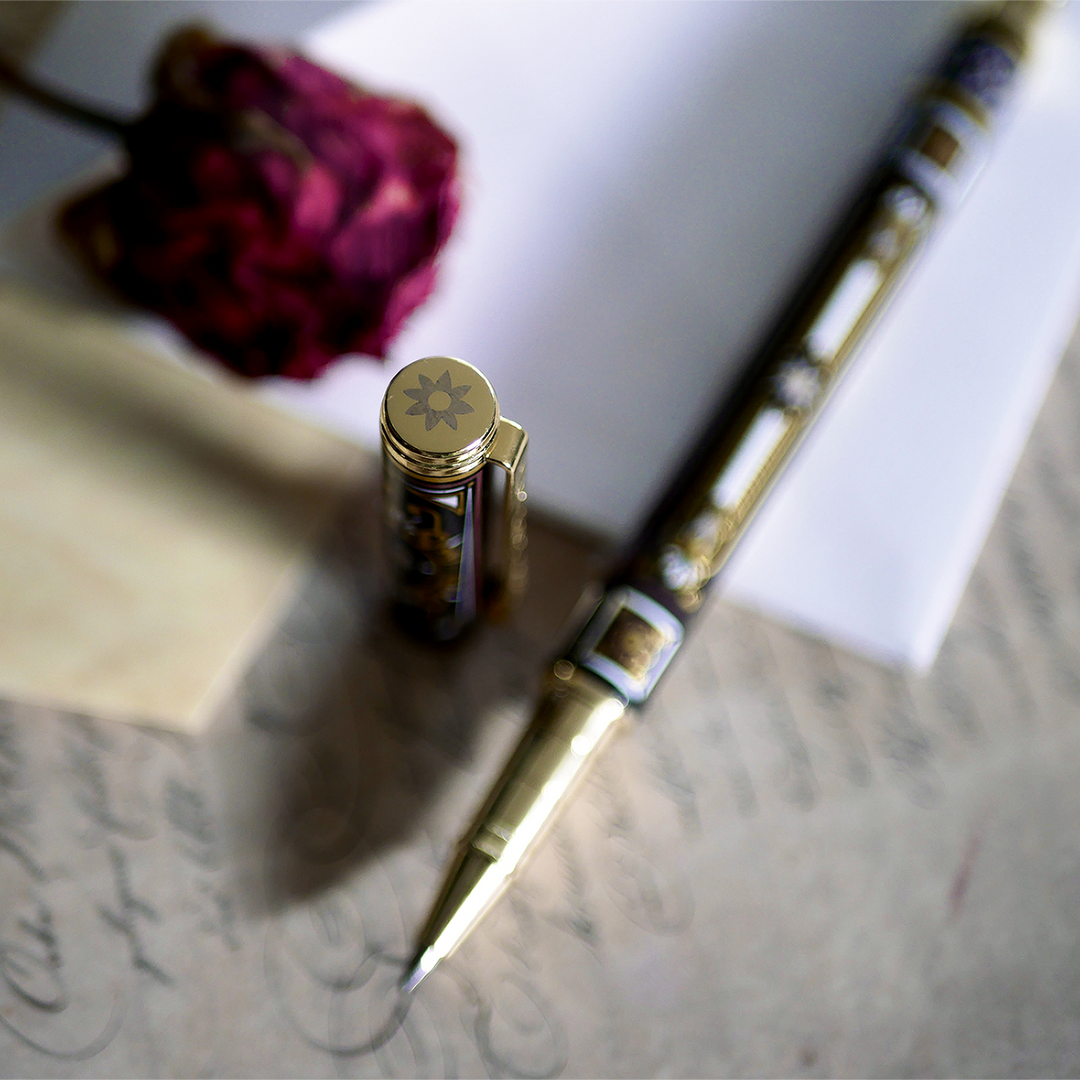 A hand holding the Black You Got This Pen by The Quirky Cup Collective, showcasing its intricate gold and white floral and geometric barrel design on a black background. The pen is positioned on top of a group of letters and envelopes with dried red roses creating a moody, vintage-inspired scene. 