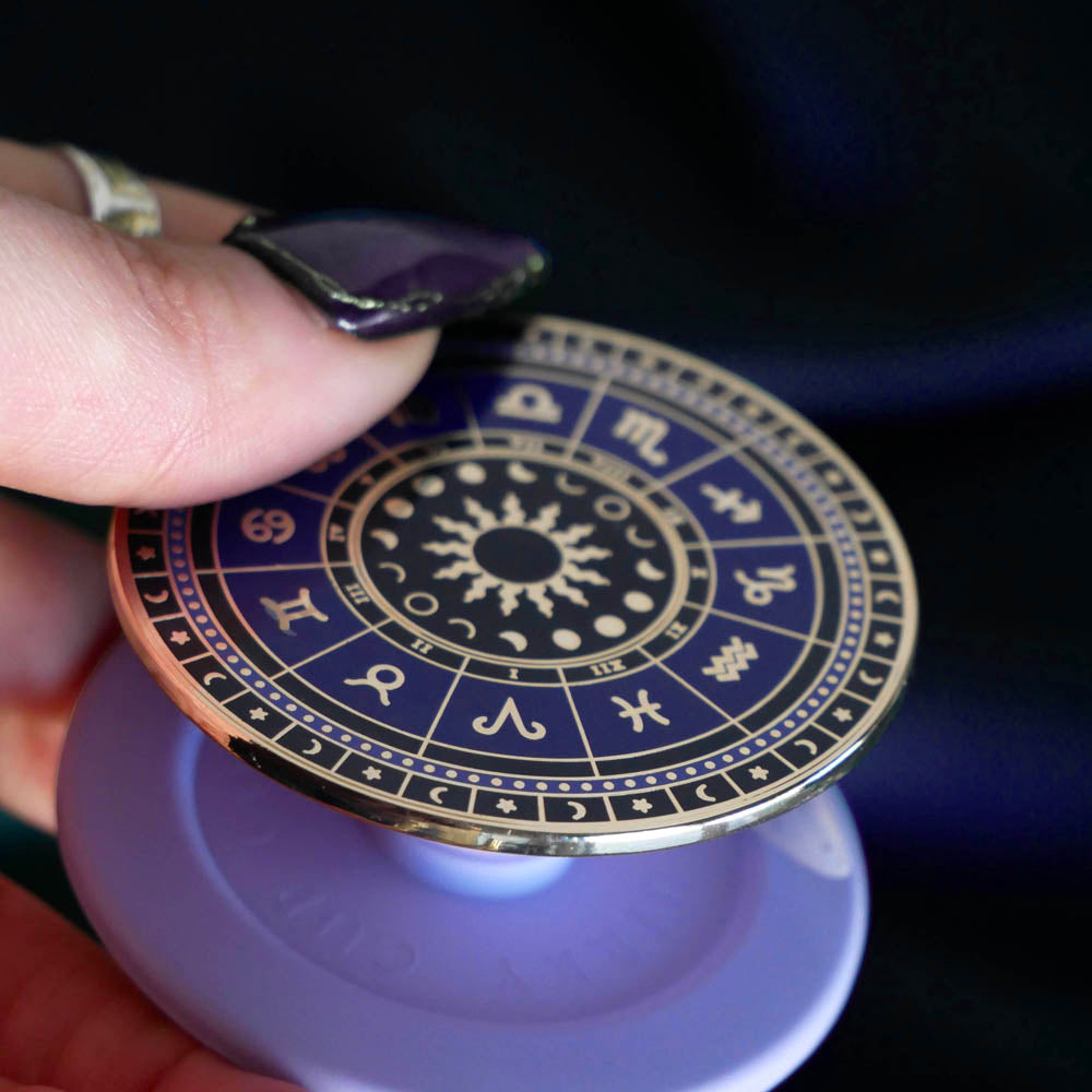 A close up detail of the zodiac clingie (think an enamel pop socket!). The kindle grip features purple and black details of the zodiac symbols around a central sun and ring of moon phases with tiny stars and moons around the edge. The plastic bit that attached to the device is a soft lilac purple.