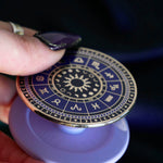 A close up detail of the zodiac clingie (think an enamel pop socket!). The kindle grip features purple and black details of the zodiac symbols around a central sun and ring of moon phases with tiny stars and moons around the edge. The plastic bit that attached to the device is a soft lilac purple.