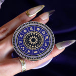 On a deep purple fabric background a hand holds the Zodiac clingie (think an enamel pop socket!). The kindle grip features purple and black details of the zodiac symbols around a central sun and ring of moon phases with tiny stars and moons around the edge.