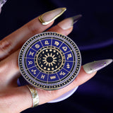 On a deep purple fabric background a hand holds the Zodiac clingie (think an enamel pop socket!). The kindle grip features purple and black details of the zodiac symbols around a central sun and ring of moon phases with tiny stars and moons around the edge.
