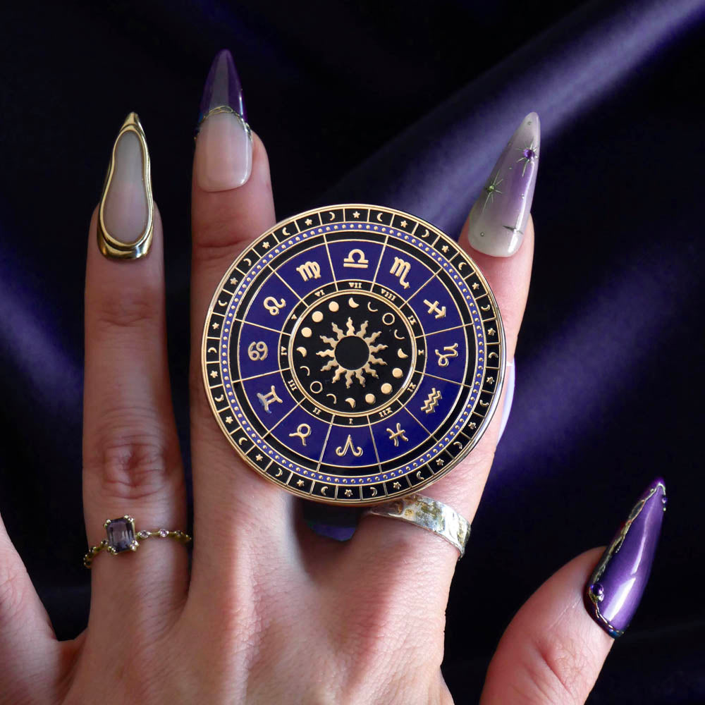 On a deep purple fabric background a hand holds the Zodiac clingie (think an enamel pop socket!). The kindle grip features purple and black details of the zodiac symbols around a central sun and ring of moon phases with tiny stars and moons around the edge.