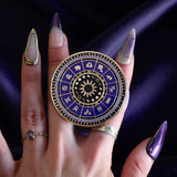 On a deep purple fabric background a hand holds the Zodiac clingie (think an enamel pop socket!). The kindle grip features purple and black details of the zodiac symbols around a central sun and ring of moon phases with tiny stars and moons around the edge.