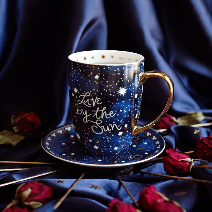 Live by the Sun Coffee Mug - Blue Mug with Gold handle and metallic gold details - Moon and Stars - Celestial Design - Le Soleil Le Gobelet - Sun Mug against satin backdrop with dried red roses and gold accessories The Quirky Cup Collective